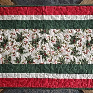 May include: A festive Christmas table runner. The runner has a central panel with a holly and berry pattern, framed by bands of red, white, and green. The quilted design adds texture and a seasonal feel to the table setting.