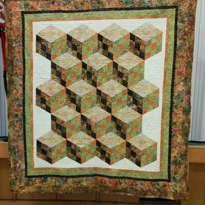 Może przedstawiać: Kołdra z geometrycznym wzorem sześcianów w odcieniach brązu, zieleni i bieli. Sześciany są ułożone w powtarzający się wzór, tworząc iluzję optyczną głębi.