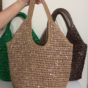 May include: A beige crochet tote bag with a grid pattern and scattered sequins, featuring two integrated handles. Behind it are two additional tote bags in the same style, one in a vibrant green and another in a dark brown, both with sequins.