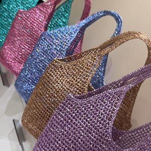 Könnte beinhalten: Eine Ansammlung von fünf Tragetaschen, die in einer Reihe angeordnet sind, jede in einer anderen Farbe wie Türkis, Pink, Blau, Gold und Violett. Diese Taschen bestehen aus einem schimmernden, grob gestrickten Netzmaterial mit einer texturierten Paillettenoberfläche.
