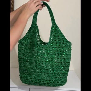 Puede incluir: Un bolso tote de color verde esmeralda vibrante hecho de material de ganchillo con un diseño de tejido abierto. El bolso está adornado con pequeñas lentejuelas verdes reflectantes integradas en el hilo, creando una textura brillante.