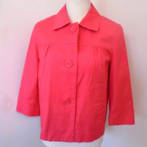 May include: A bright coral-colored jacket featuring a classic collar, three-quarter length sleeves, and a front button closure with three matching buttons. The garment includes subtle gathered detailing at the chest area, providing a structured fit.