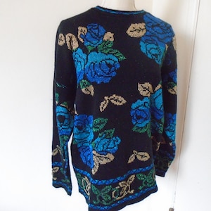 May include: A black long-sleeve sweater featuring a knit pattern of large blue roses with green leaves and gold accents. The garment has a crew neckline and a decorative horizontal band with blue and green geometric patterns along the bottom hem.