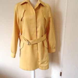 May include: A bright yellow trench coat featuring a pointed collar, long sleeves with buttoned cuffs, and a matching belt with a silver-toned metal D-ring buckle. The coat includes two chest pockets with button flaps and a concealed front button placket.