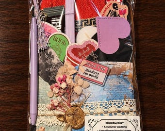 May include: A clear plastic gift bag containing a pair of red knit socks, a lavender pen, a heart-shaped purple bookmark, a small dried flower bouquet with a gold wax seal, and a card labeled ROMCOM/COZY with book-themed stickers and a keychain.