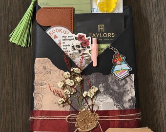 May include: A black paper-wrapped book gift set featuring a red ribbon, a gold wax seal, dried flowers, a green tassel bookmark, a Taylors tea bag, a yellow honey lollipop, a skull sticker, a book-themed keychain, and a tag labeled The Collected Chapter.
