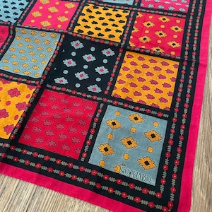 May include: A square scarf featuring a patchwork grid of various floral and geometric patterns in shades of red, yellow, black, and light blue. The fabric has a red border with a small floral print and the text SOULEIADO printed in the corner.