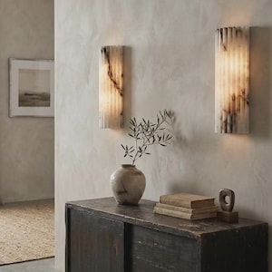 May include: Two vertical wall sconces made of white and grey veined stone with a fluted, cylindrical design. The lights emit a soft, warm glow from the top and bottom, highlighting the natural texture and patterns of the stone material.