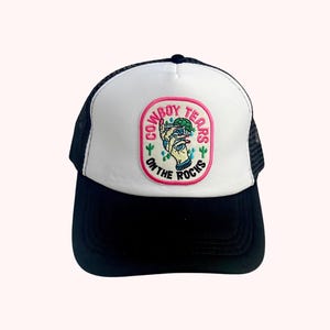 May include: A black and white trucker hat with a white front panel. The front features a patch with the text "Cowboy Tears on the Rocks" and a hand holding a drink with cacti. The hat has a black brim and mesh back.