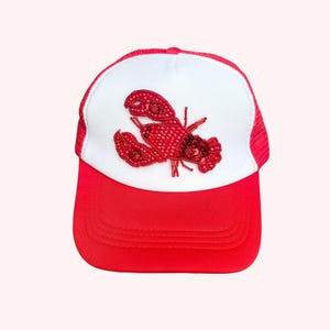 May include: Red and white trucker hat with a red lobster design. The hat has a red peak and mesh back, with a white front panel. The lobster is embellished with red sequins and beads, adding a touch of sparkle to the design.