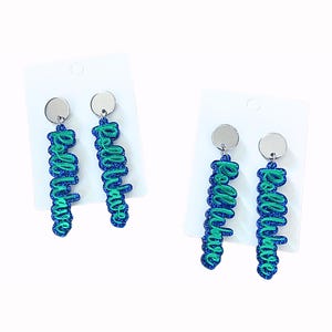 May include: A pair of dangling earrings with the word "Real Babe" in a cursive font. The earrings are blue and green glitter with a silver disc at the top. The earrings are displayed on white earring cards.