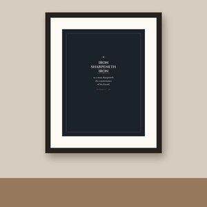 May include: A framed wall art print featuring a dark navy blue background with white serif text that reads: IRON SHARPENETH IRON so a man sharpeneth the countenance of his friend. PROVERBS 27:17. The print is encased in a simple black frame.