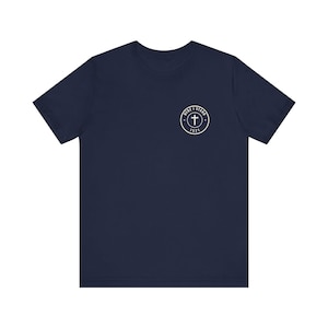 May include: A navy blue short-sleeve t-shirt featuring a small white circular graphic on the left chest. The graphic contains a cross symbol in the center, surrounded by the text "HERE I STAND" at the top and the number "1521" at the bottom.