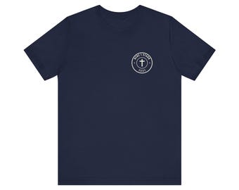May include: A navy blue short-sleeve t-shirt featuring a small white circular graphic on the left chest. The graphic contains a cross symbol in the center, surrounded by the text "HERE I STAND" at the top and the number "1521" at the bottom.