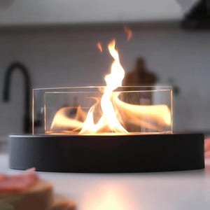 May include: A tabletop fireplace featuring a circular black base and a clear glass cylindrical guard. Bright orange and yellow flames rise from the center of the unit, creating a visible fire effect within the transparent glass enclosure.