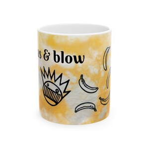 Può includere: Una tazza in ceramica bianca con motivo tie-dye giallo, decorata con disegni a linee nere di banane e un volto con capelli a punta. Il testo "as & blow" è stampato in nero sul lato della tazza, che presenta una finitura liscia.