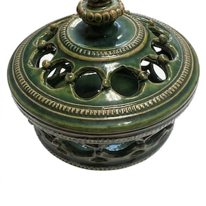 May include: A round, dark green glazed ceramic jar with a matching lid. The lid features a decorative knob and cut-out openings. The body of the jar also has a series of cut-out openings around the perimeter, creating a lattice-like appearance.
