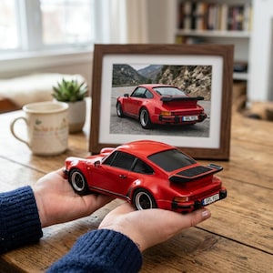 Op de afbeelding: Een rood schaalmodel van een sportauto met een zwarte achterspoiler en zwarte wielen, voorzien van een kentekenplaat met de tekst GRL 911. Het model wordt in de handen gehouden en weerspiegelt de rode sportauto op de ingelijste foto.