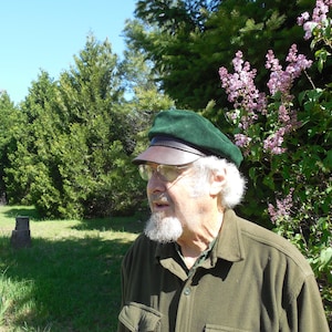 May include: An older person wearing a green cap with a brown brim and glasses, and a green button-up shirt. The person has a white beard and is standing outside in front of green trees and purple flowers.