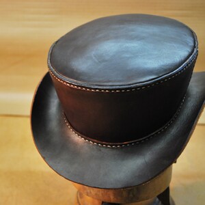 May include: A dark brown leather top hat with a rounded crown and a wide brim. The hat features visible stitching in a lighter color along the seams. The hat is displayed on a wooden form.
