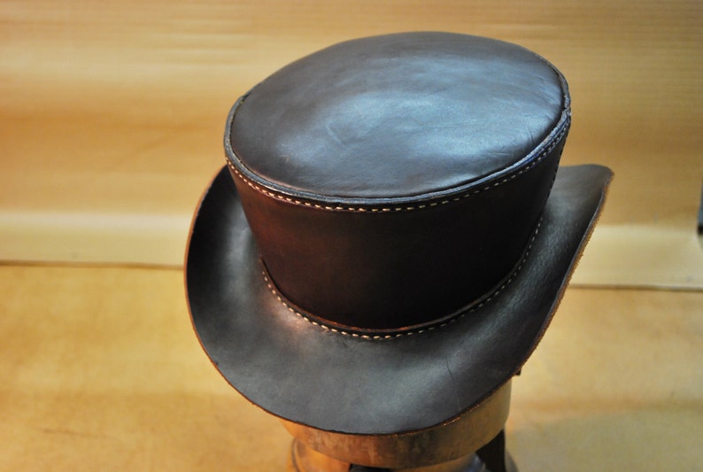 May include: A dark brown leather top hat with a rounded crown and a wide brim. The hat features visible stitching in a lighter color along the seams. The hat is displayed on a wooden form.