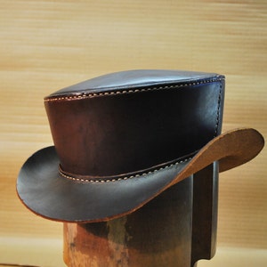 May include: A dark brown leather top hat with a wide brim. The hat has white stitching along the brim and the top. The hat is displayed on a wooden hat stand. The hat is approximately 6 inches tall.
