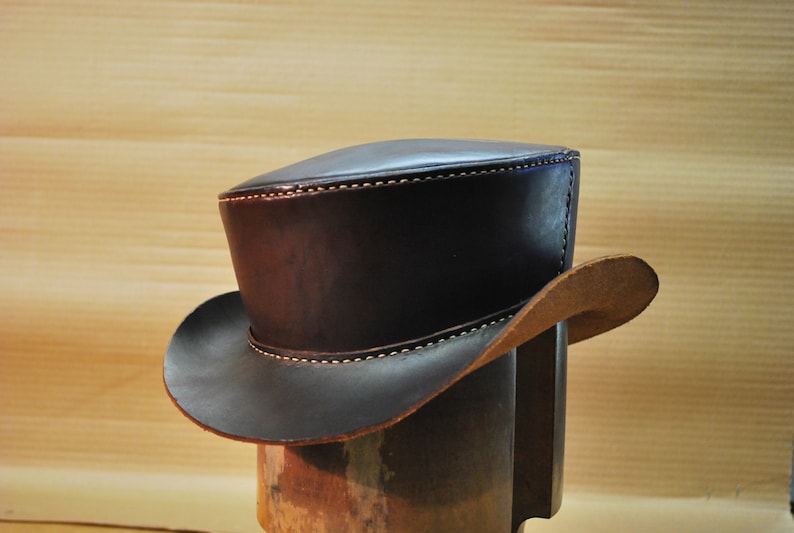 May include: A dark brown leather top hat with a wide brim. The hat has white stitching along the brim and the top. The hat is displayed on a wooden hat stand. The hat is approximately 6 inches tall.