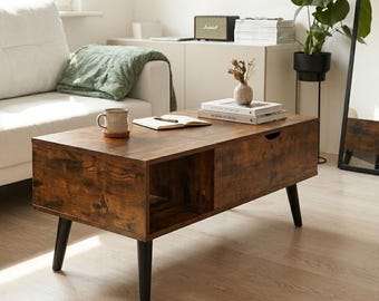 May include: A rectangular coffee table with a dark brown wood grain finish, featuring four tapered black legs, an open side storage compartment, and a lift-top surface. The table holds a ceramic mug, an open notebook, and two stacked books.