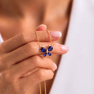 May include: A gold necklace featuring a butterfly-shaped pendant. The pendant is composed of four pear-cut blue stones forming the wings, accented with two small clear stones at the top, suspended from a delicate gold chain.