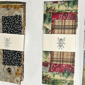 May include: Four sets of folded fabric wraps secured with white paper bands featuring a bee illustration. The wraps display various patterns including green polka dots, yellow sunflowers, black leaf prints, red paisley, brown plaid, and forest scenes with bears.