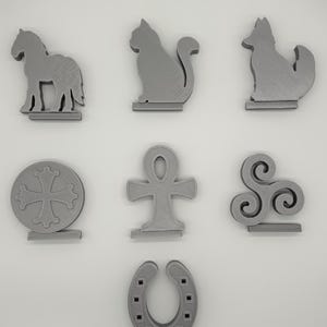 Peut inclure: Un ensemble de sept figurines grises imprimées en 3D. La collection comprend les silhouettes d'un cheval, d'un chat, d'un loup, d'une croix circulaire, d'un symbole ankh, d'un triskèle à triple spirale et d'un fer à cheval.