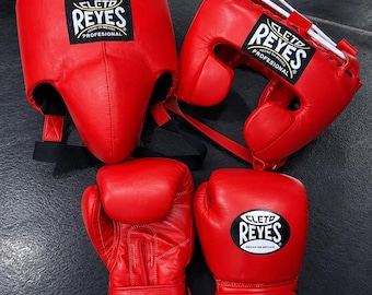 May include: A set of bright red boxing equipment including a headgear piece and a pair of boxing gloves. Each item features the black and white Cleto Reyes Profesional logo patch. The headgear has white lace adjustments and the gloves have wrist straps.