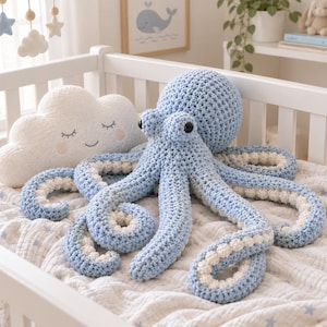 May include: A large, crocheted octopus plush toy with a light blue exterior and white underside on its eight tentacles. The octopus features two round black eyes and a textured, knotted yarn construction. Beside it sits a white cloud-shaped pillow.