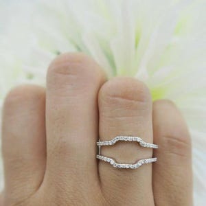 Peut inclure: Une bague protectrice argentée composée de deux anneaux parallèles reliés sur les côtés, chacun incrusté d'une rangée de petites pierres claires et scintillantes. Le design crée un espace ouvert au centre pour encadrer une autre bague.