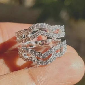 May include: A silver-toned ring guard featuring two curved bands connected by a bridge. Both bands are encrusted with small, clear, round-cut stones that cover the entire surface, creating a continuous sparkling effect across the metal.