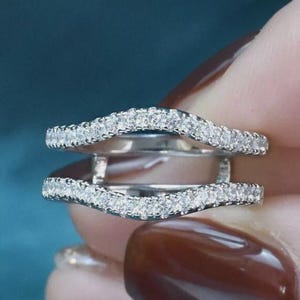 May include: A silver-toned ring guard featuring two curved bands connected by a central bridge. Both bands are encrusted with a continuous row of small, clear, sparkling stones that follow the wavy, contoured shape of the metal frame.