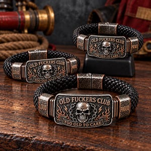 Puede incluir: Tres pulseras de cuero trenzado negro con una placa metálica rectangular que muestra un gráfico de calavera, detalles de cadenas y llamas. El texto dice OLD FUCKERS CLUB y TOO OLD TO CARE, con LOUD y PROUD grabados en los laterales.