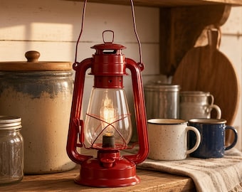 Puede incluir: Un farol de tormenta de metal rojo brillante con una chimenea de vidrio transparente y un asa de alambre. El farol cuenta con una bombilla central con filamento visible, una base circular y una jaula de alambre protectora.