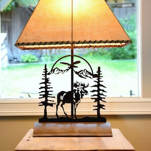 May include: A table lamp with a beige lampshade and a metal base featuring a silhouette of a moose and pine trees. The lamp is on a wooden table.