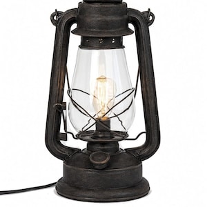 Pu&ograve; includere: Una lanterna in metallo color bronzo scuro con un globo in vetro trasparente e una lampadina a filamento interna. La lanterna presenta una maniglia superiore, una base circolare e un cavo di alimentazione nero. Testo: C UL US LISTED e DIMMABLE.