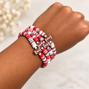 May include: A set of four stacked bracelets made of small, flat, circular clay beads in alternating red, white, and grey colors. Each bracelet features a brown, oval-shaped charm designed to look like an American football with white laces.