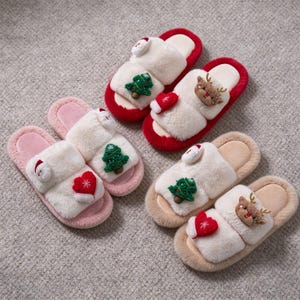 Puede incluir: Dos pares de pantuflas abiertas de felpa con tiras de piel sintética blanca, decoradas con aplicaciones bordadas de árboles de Navidad, renos, gorros de Papá Noel y corazones. Un par tiene suelas rojas y el otro suelas rosa claro y beige.