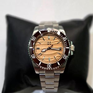 May include: A stainless steel wristwatch featuring a metallic copper-toned dial with a horizontal wave pattern, a dark maroon rotating bezel with white numerals, and a silver-toned link bracelet with a central logo at the twelve o'clock position.