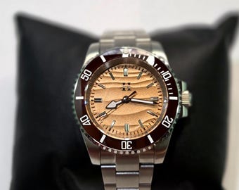 May include: A stainless steel wristwatch featuring a metallic copper-toned dial with a horizontal wave pattern, a dark maroon rotating bezel with white numerals, and a silver-toned link bracelet with a central logo at the twelve o'clock position.