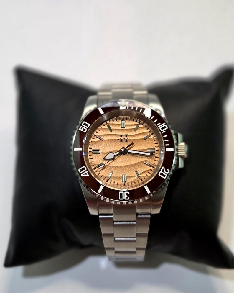 May include: A stainless steel wristwatch featuring a metallic copper-toned dial with a horizontal wave pattern, a dark maroon rotating bezel with white numerals, and a silver-toned link bracelet with a central logo at the twelve o'clock position.