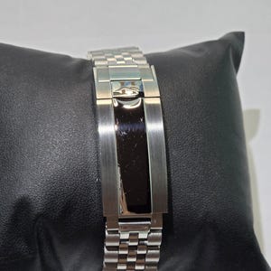 May include: A silver-tone metal watch bracelet featuring a multi-link design and a central deployment clasp. The clasp has a polished black rectangular center panel and a small engraved logo at the top, showcasing a refined metallic finish.