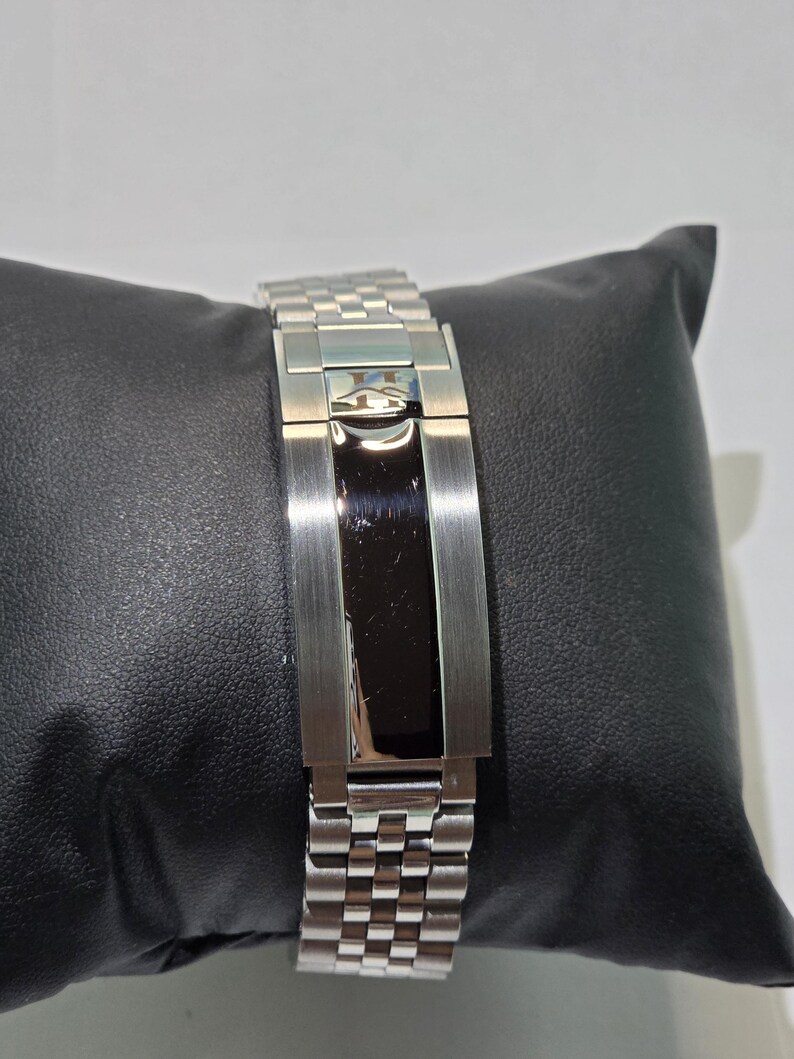 May include: A silver-tone metal watch bracelet featuring a multi-link design and a central deployment clasp. The clasp has a polished black rectangular center panel and a small engraved logo at the top, showcasing a refined metallic finish.