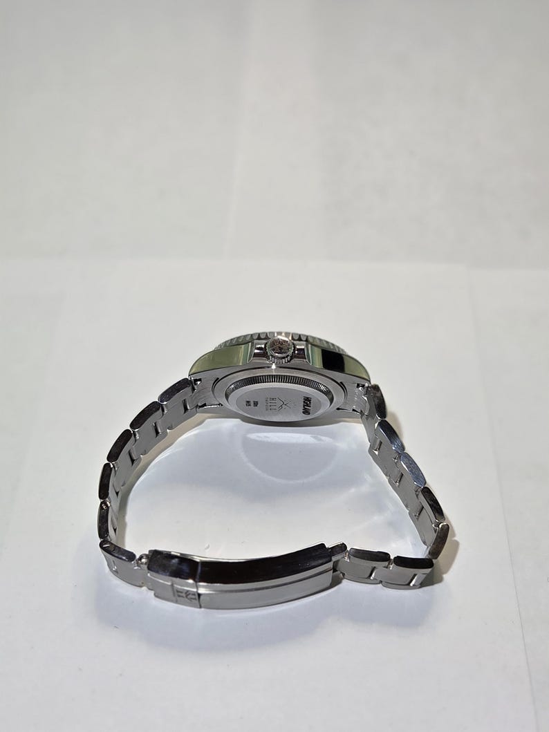 May include: A silver-toned stainless steel wristwatch with a metal link bracelet. The back of the watch case features engraved text that reads HCLAND and HILL, along with a small logo. The watch is shown from an angled perspective.