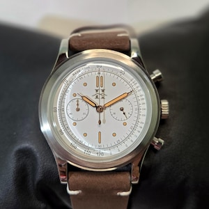 May include: A silver-tone chronograph wristwatch with a white dial, copper-colored hands, and indices. The watch features a tachymeter scale, two sub-dials, and a brown leather strap with white stitching. A logo is printed at the twelve o'clock position.