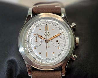 May include: A silver-tone chronograph wristwatch with a white dial, copper-colored hands, and indices. The watch features a tachymeter scale, two sub-dials, and a brown leather strap with white stitching. A logo is printed at the twelve o'clock position.